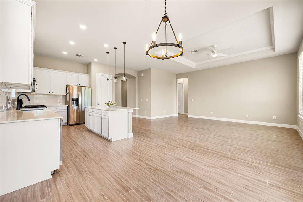 View from breakfast nook showcases the spacious open-plan kitchen and living area with modern fixtures. The kitchen features white cabinetry, stainless steel appliances, and an island with pendant lighting.