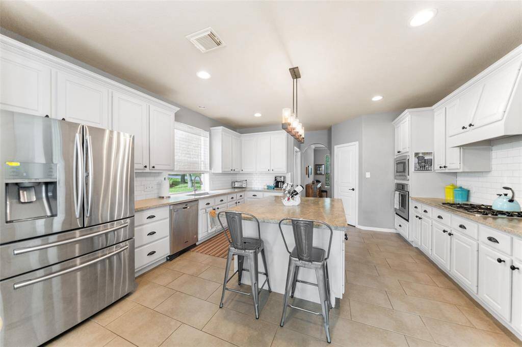 This kitchen features bright, modern white cabinetry and countertops with a spacious island. It includes stainless steel appliances, a gas cooktop, and stylish lighting, creating a welcoming and functional cooking space.