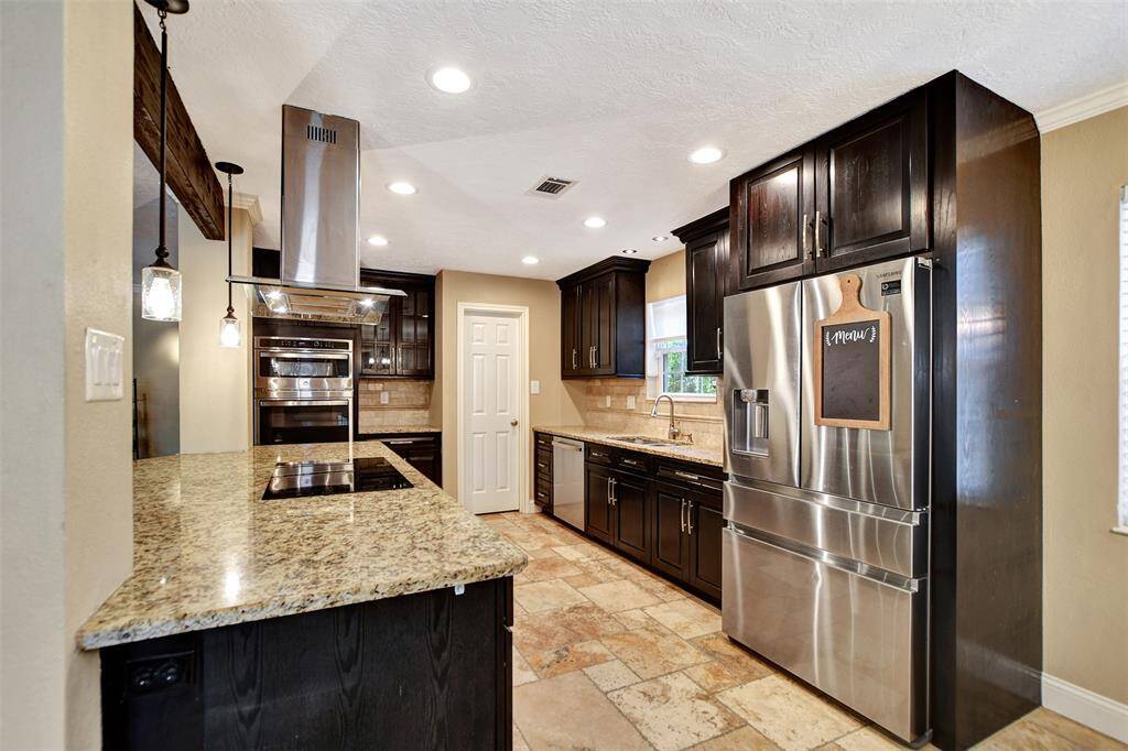 Modern kitchen with granite countertops and stainless steel appliances. Features dark wood cabinetry, a built-in oven, and a sleek hood vent. Ample lighting and tiled flooring complete the sophisticated look.