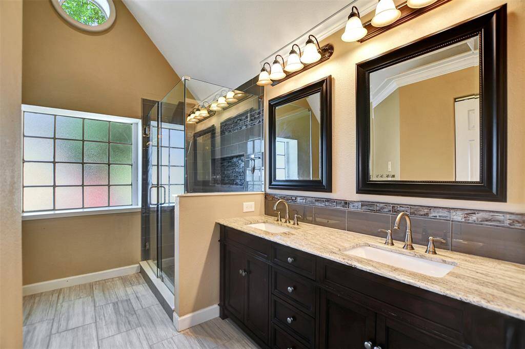 Primary  bathroom features a sleek double vanity with granite countertops, modern fixtures, and framed mirrors. It includes a spacious glass-enclosed shower and abundant natural light from large frosted windows, creating a bright and inviting space.