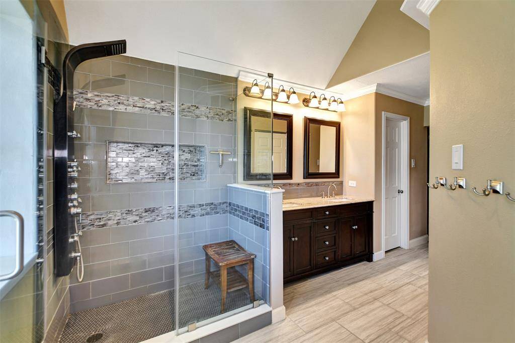 Spacious glass-enclosed shower with modern tile accents and multiple showerheads. It includes a double vanity with a dark wood cabinet, large mirrors, and stylish lighting.