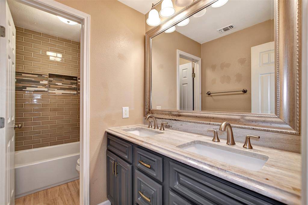Upstairs bath offers modern double vanity with sleek faucets and a large framed mirror. The shower area has stylish beige brick tiles with a decorative accent strip.