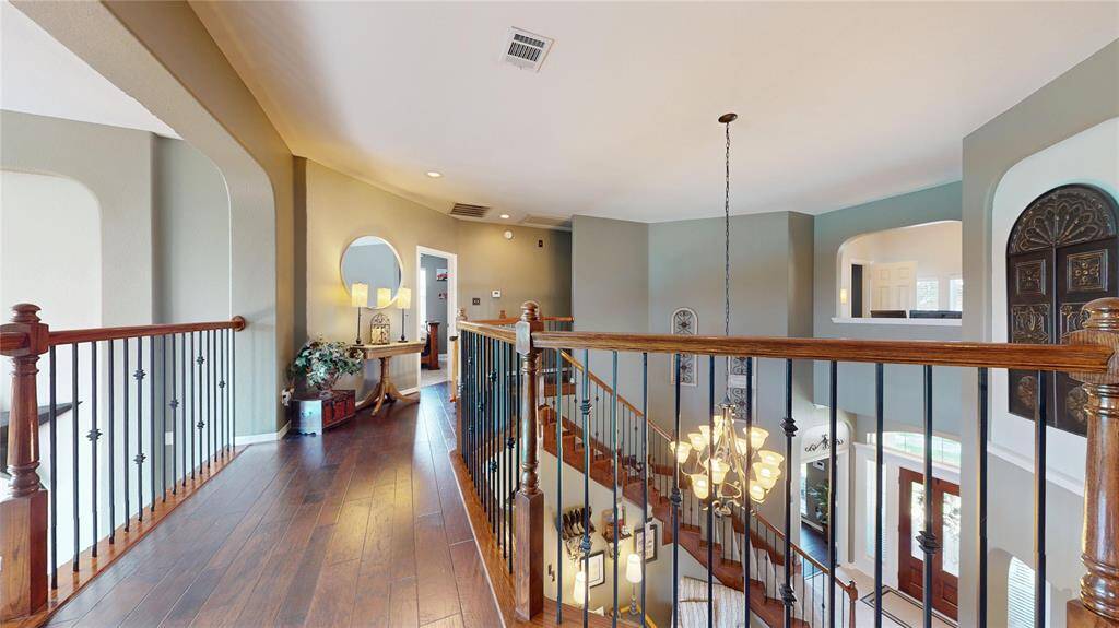 The upstairs landing offers a beautiful transition space with rich hardwood flooring and elegant wrought-iron railing that overlooks the grand entry and living area below. Arched openings and custom wall niches add architectural interest, while recessed lighting enhances the warm, inviting ambiance. This versatile space is ideal for a reading nook, decorative display, or simply enjoying the view of the chandelier and soaring ceilings below.