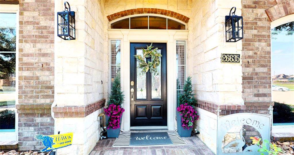 Step into timeless elegance with this beautifully designed entryway featuring a classic brick and stone façade. The rich, dark front door with decorative glass insets is framed by transom and sidelight windows, allowing natural light to enhance the warm welcome. Flanked by neatly trimmed evergreens and vibrant seasonal flowers, the covered porch adds a cozy, polished touch. Stylish lantern sconces and a welcoming doormat complete the picture-perfect entrance.