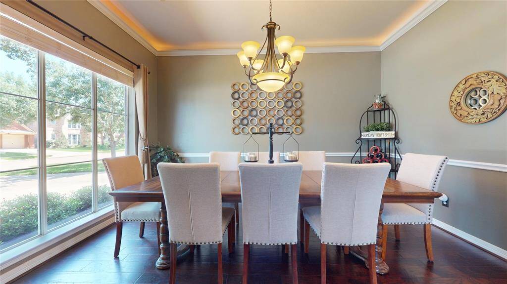 This sophisticated formal dining room offers a warm and welcoming space ideal for entertaining. Anchored by rich wood flooring and crowned with classic wainscoting and crown molding, the room exudes timeless elegance. A large picture window fills the space with natural light and provides lovely views of the front yard, while a graceful chandelier and ambient cove lighting add a refined glow to evening gatherings.