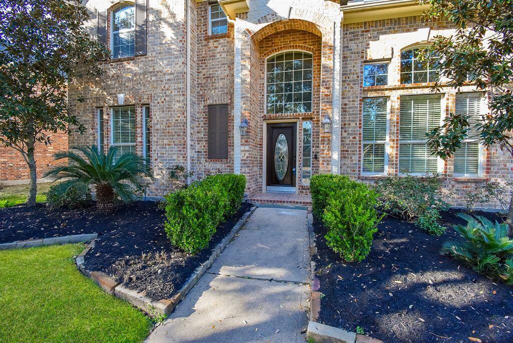 Front exterior of a brick home with a welcoming arched doorway and paved walkway is surrounded by well-maintained shrubs and mature trees.