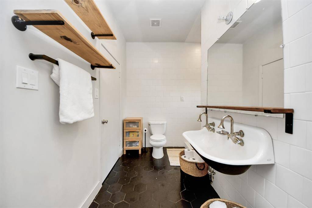 This bathroom includes a double basin sink with industrial-style faucets, wooden shelving, and an oversized mirror. The space is well-lit and has a minimalist and modern aesthetic.