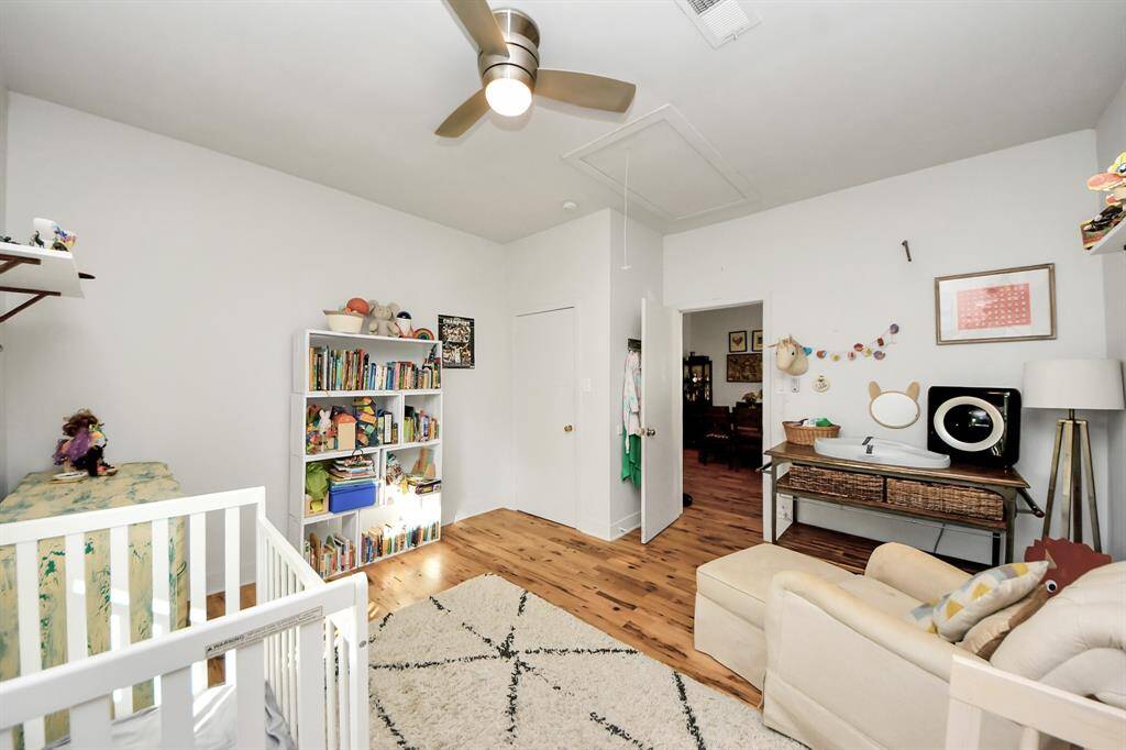This is a bright, cozy nursery with hardwood floors. There's ample storage with open shelving for books and toys, and a ceiling fan for comfort. The room is well-lit with natural light from the window.
