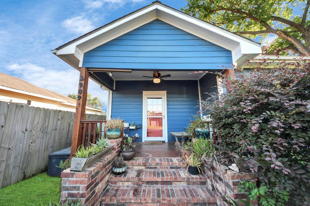 Charming blue HardiePlank bungalow-cottage home, with a welcoming front porch. Features brick steps, lush plants, and a cozy seating area, perfect for relaxing. Enclosed by a iron fence, offering privacy and character.