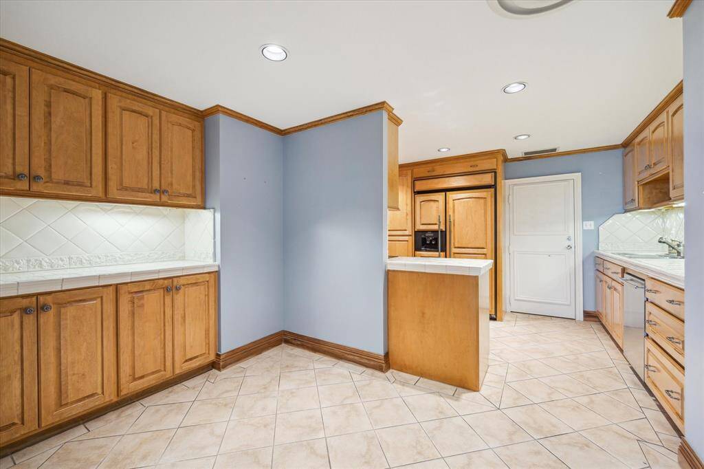 Kitchen with tile flooring, tile countertops/backsplash, custom cabinetry, & recessed lighting. Back door leading to hallway (convenient access for groceries & trash).