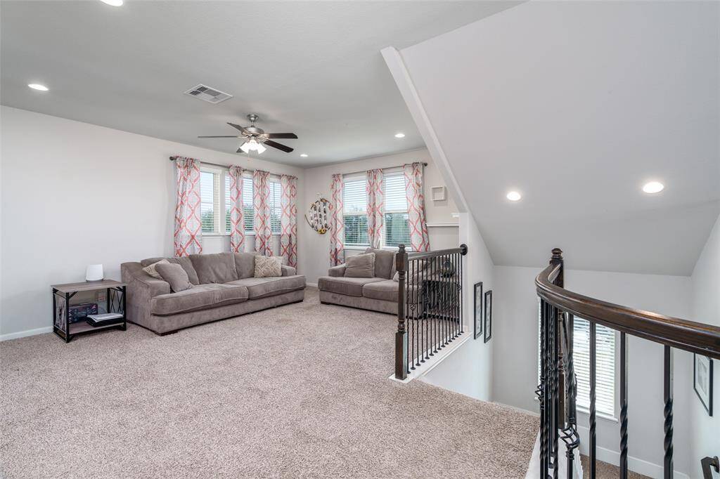 The upstairs game room or additional family room offers a cozy and versatile space, featuring plush carpet, a ceiling fan, recessed lighting, and multiple windows with privacy blinds that fill the room with natural light.