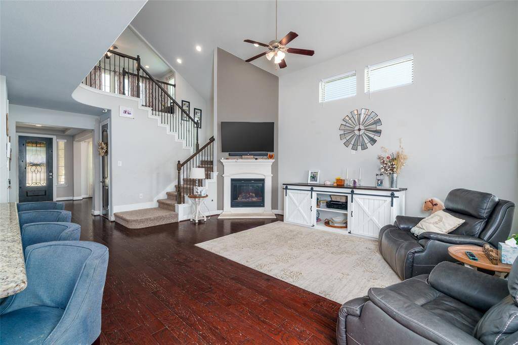 The inviting living room showcases engineered wood flooring, a soaring high ceiling, a cozy gas log fireplace, and a wall of windows with privacy blinds that fill the space with natural light.
