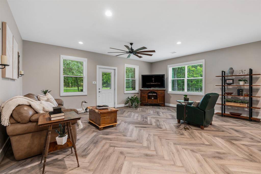 This is a spacious, well-lit living room with modern herringbone wood plank flooring, neutral wall colors, and ample natural light from large windows. It features a comfortable seating area, a ceiling fan, and a designated space for entertainment equipment. The room also provides views and direct access to the outdoors and a large rear porch.