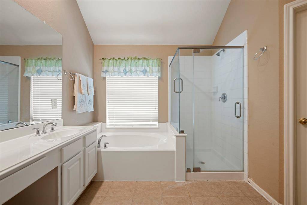 This bright bathroom features a large soaking tub, a glass-enclosed shower, dual sinks with a white countertop, and a window with decorative valances, offering a clean and inviting space.