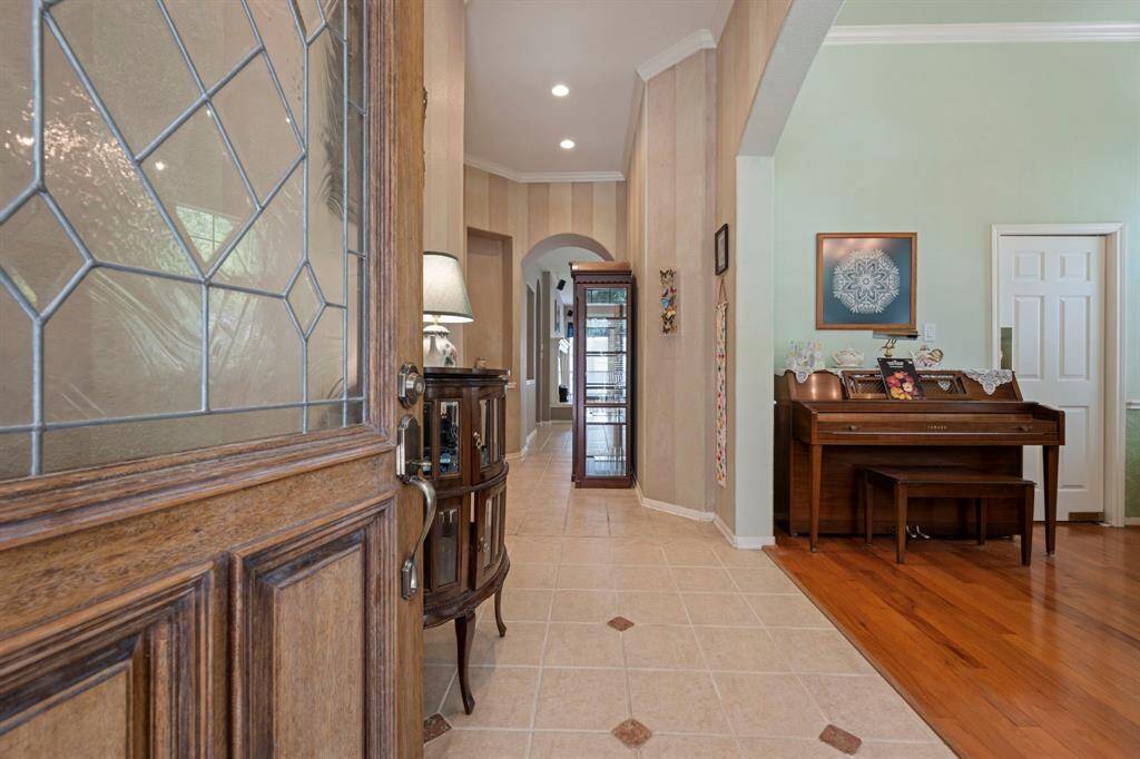 This inviting entryway features a wooden door with decorative glass, leading into a spacious hallway with tiled flooring. The area has warm, neutral tones, crown molding, and a mix of tile and hardwood floors creating a welcoming and elegant atmosphere.