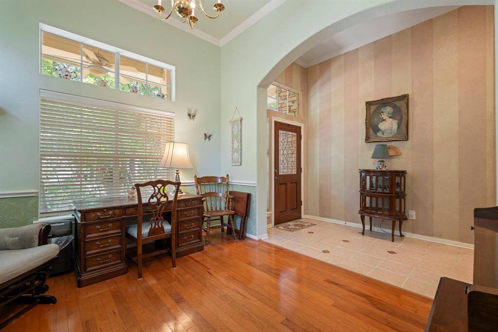 This inviting entryway features warm wood flooring, ample natural light from large windows, and elegant touches like a chandelier and decorative wall accents. The space is currently accentuated by a cozy desk area, and is perfect for a small office or reading nook.