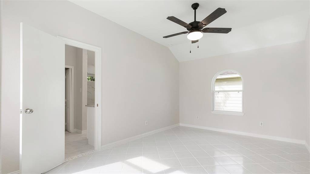 This bright, airy room features a high ceiling with a modern ceiling fan, neutral walls, and a tiled floor. A charming arched window allows natural light to enter, and there's an adjacent bathroom visible through an open door.