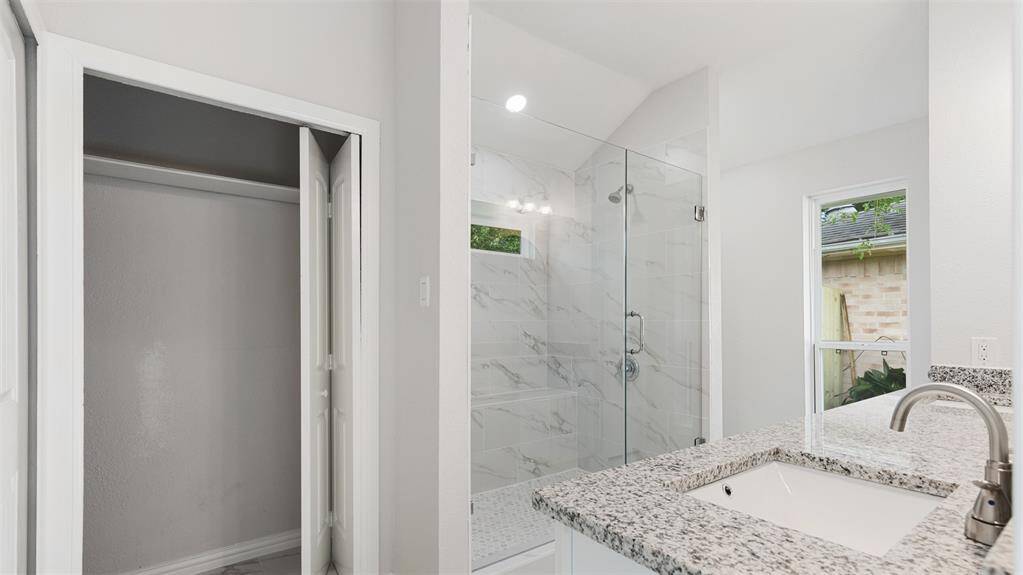 This modern bathroom features a sleek design with a glass-enclosed shower, marble-like tile, and a granite countertop. It includes a spacious sink area and a small window for natural light.