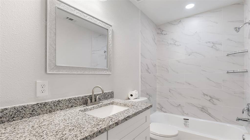 This bathroom features a sleek design with a granite countertop, under-mount sink, and a modern faucet. The walls and bathtub area are adorned with elegant marble-style tiles. A large framed mirror and recessed lighting add a bright, sophisticated touch.