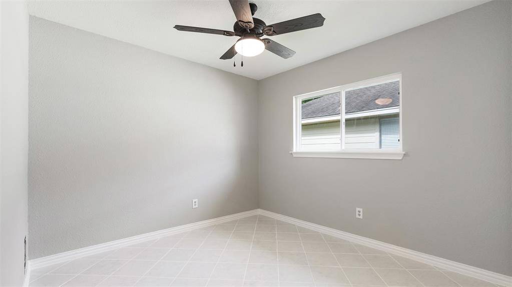 This room features neutral gray walls, a ceiling fan with a light, a window for natural light, and light-colored tiled flooring, offering a clean and versatile space.