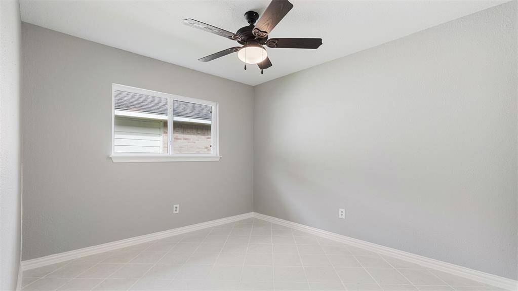 This room is bright and airy with light gray walls and a white tile floor. It features a ceiling fan with a central light and a window that lets in natural light. Perfect for a small bedroom or office.
