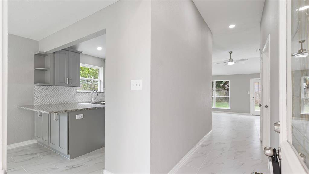 Bright and modern interior with light gray walls and tile flooring. Features a kitchen with granite countertops and herringbone backsplash, leading to an open living area with large windows and a ceiling fan.