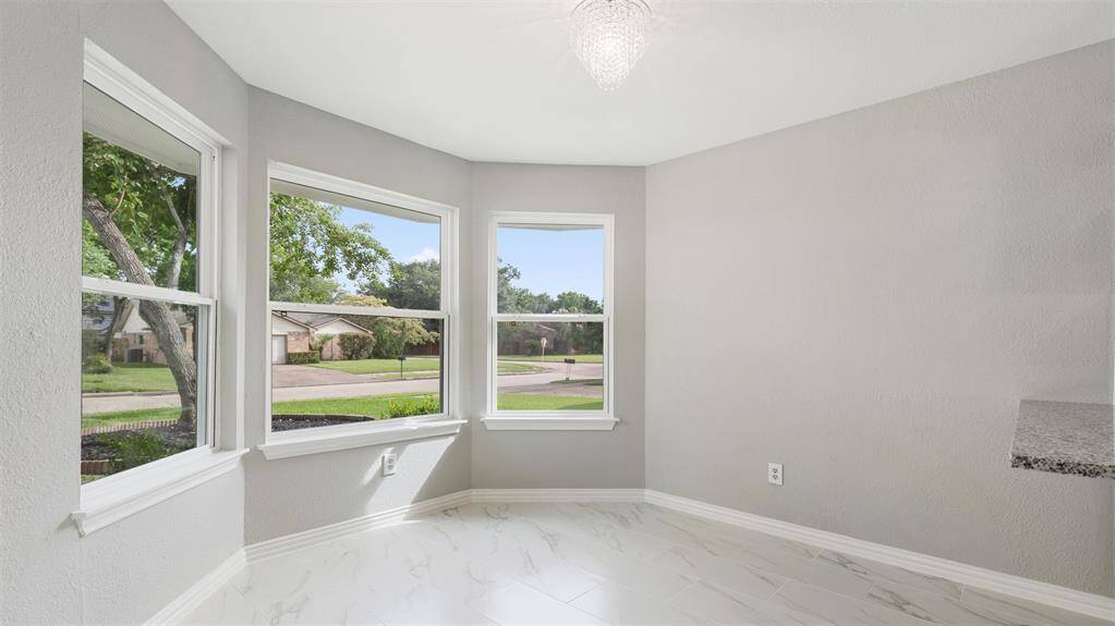 Bright and inviting corner room with large windows offering a view of a suburban street. It features light gray walls, marble-patterned flooring, and a small chandelier, perfect for a cozy breakfast nook or reading area.