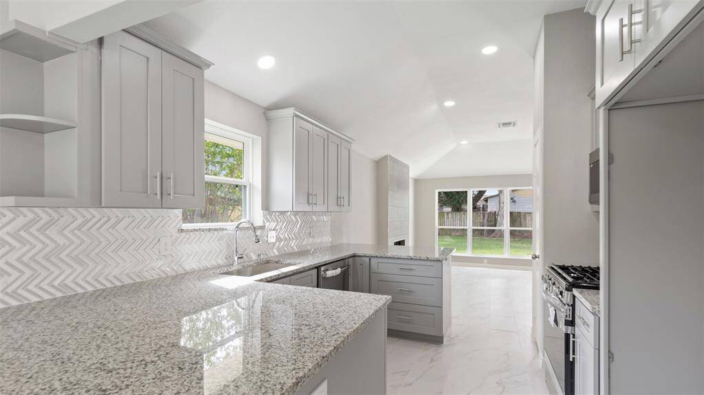 This kitchen features sleek gray cabinetry, granite countertops, and a stylish herringbone backsplash. It opens to a bright living area with large windows overlooking the backyard, creating a spacious and modern feel.