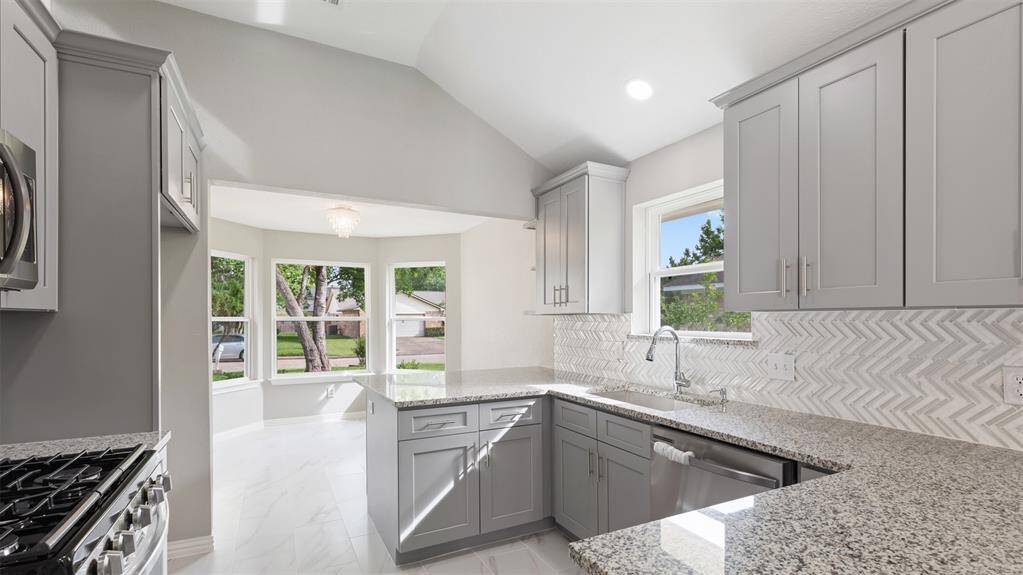 This bright kitchen features modern gray cabinetry, granite countertops, and a stylish herringbone backsplash. It offers a cozy breakfast nook with large windows, allowing plenty of natural light and a view of the outdoors.
