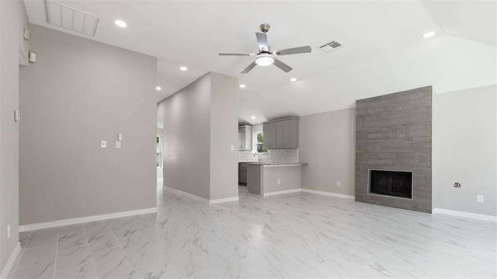 This modern living space features sleek tile flooring, neutral gray walls, and recessed lighting. The room includes a contemporary ceiling fan and a stylish, tiled fireplace. The open layout leads to a kitchen area with gray cabinetry and a granite countertop.