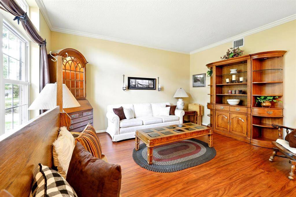 Step into the elegant formal living room featuring rich wood floors and classic crown molding that add timeless character to the space. A large picture window, topped with a transom window, fills the room with natural light.