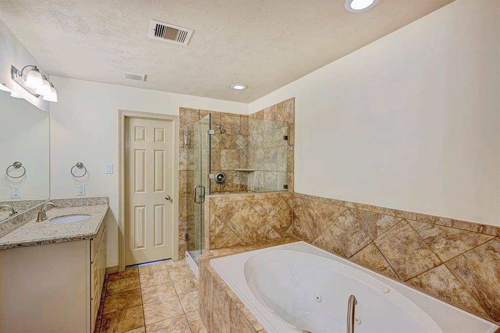 The primary bathroom features a spacious design with a neutral color palette. It includes a large built-in soaking tub with a brushed metal faucet, surrounded by elegant beige and tan ceramic tiles. Adjacent to the tub is a glass-enclosed walk-in shower with matching tiled walls and a built-in corner shelf, providing a sleek and cohesive look.