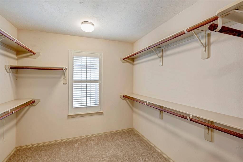 The primary closet is a spacious walk-in design featuring multiple wooden shelves and hanging rods, providing ample storage for clothing and accessories. The room is well-lit by a ceiling-mounted light fixture, enhancing the visibility of the space. The closet's neutral-toned walls and soft carpet flooring add a cozy and functional atmosphere. A window with classic white shutters allows natural light to filter in, brightening up the area.