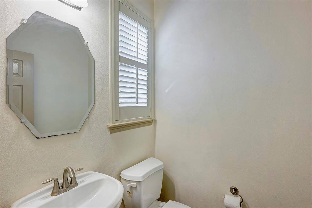 The powder room is compact and functional, featuring a pedestal sink with a brushed nickel faucet and an octagonal wall-mounted mirror that adds a unique touch. A small window with plantation shutters allows for natural light while maintaining privacy. The neutral color palette creates a bright and airy atmosphere, making the space feel welcoming and tidy.