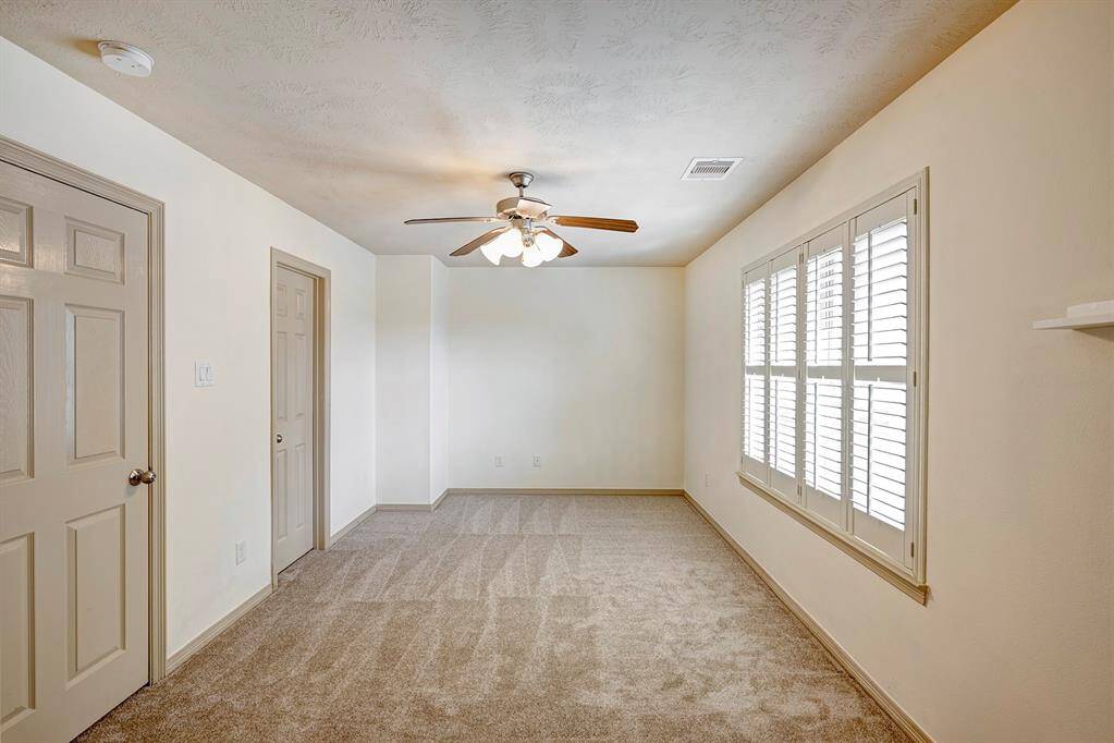 This secondary bedroom spacious and bright, featuring a soft carpet and neutral-colored walls. A ceiling fan with light fixtures adds both comfort and illumination to the room. Large windows with white plantation shutters provide plenty of natural light while offering privacy control. The room is versatile and can be used as a guest bedroom, office, or additional living space. Multiple doors lead to a closet and bathroom, enhancing functionality.