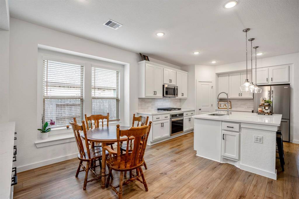 From this angle, you get a clear view of the open dining area flowing seamlessly into the kitchen. The spacious layout allows for easy movement between the two areas, while the cabinetry along the dining room wall and the kitchen’s island create a cohesive and functional design. The large windows bring in natural light, further enhancing the bright and inviting atmosphere throughout.