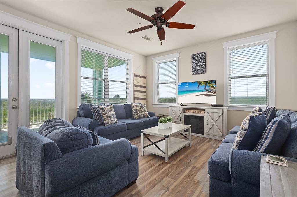 Welcome to a bright and airy living room, where comfort meets coastal charm. Large windows and French doors invite natural light, creating a warm and inviting atmosphere.