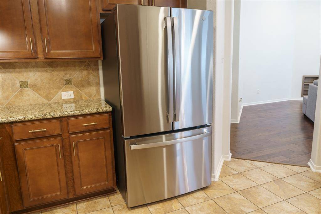 This kitchen features a stainless steel French door refrigerator set against a backdrop of warm wood cabinets and a granite countertop. The tiled floor complements the neutral tones, and the space opens into a room with hardwood flooring. Refrigerator included.