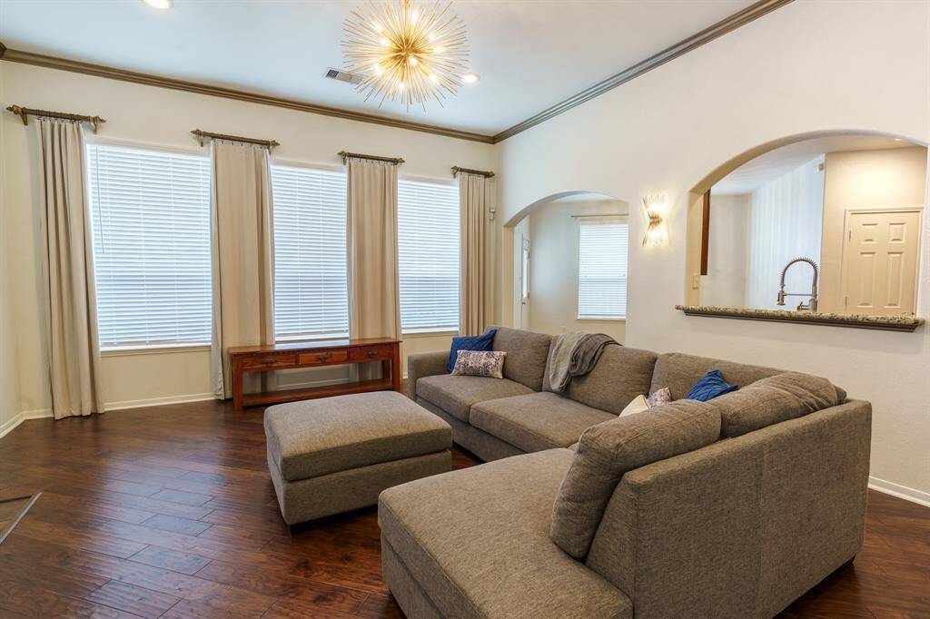 This inviting living room features  warm wood flooring. Large windows adorned with elegant drapes allow natural light to fill the space. A modern light fixture and stylish wall sconces add a contemporary touch, while arched openings provide a view into the adjacent kitchen area.