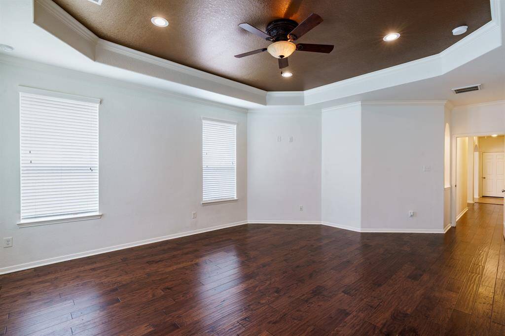 This photo showcases a Primary Bedroom  with dark wood flooring, white walls, and a stylish tray ceiling featuring recessed lighting and a ceiling fan. Four windows provide natural light, and there's an open hallway leading to other areas of the home.