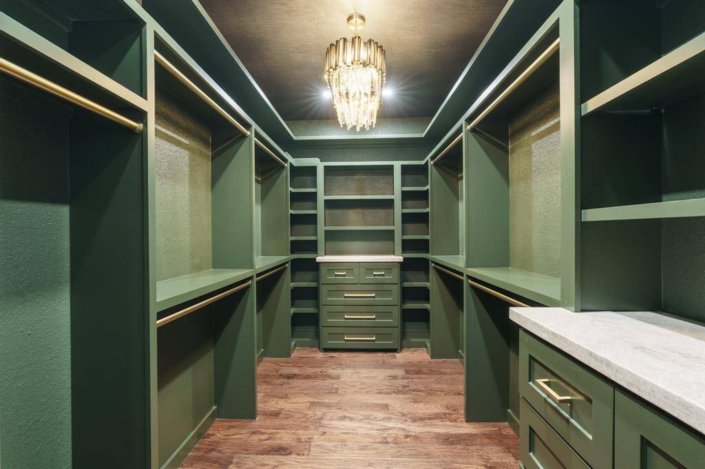 This spacious walk-in closet features elegant green cabinetry with ample shelving and hanging space. It includes a luxurious chandelier and hardwood flooring, providing a stylish and functional storage solution.