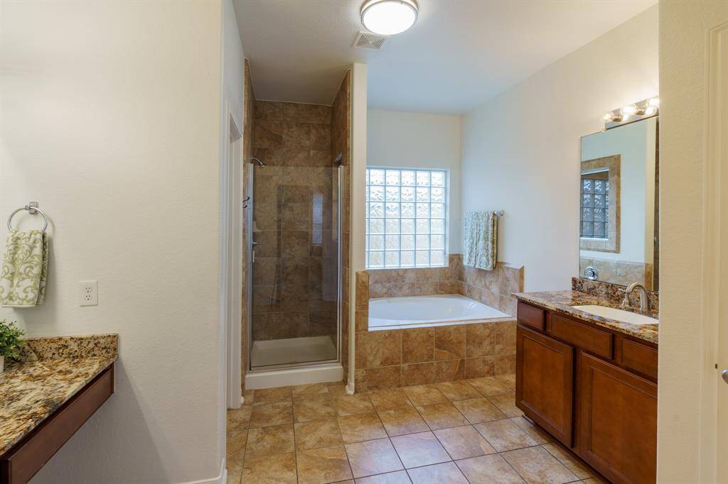 This bathroom features a spacious layout with a granite-topped vanity, a large mirror with bright lighting, and a tiled floor. It includes a built-in bathtub with tile surround and a storage closet, offering both style and functionality.