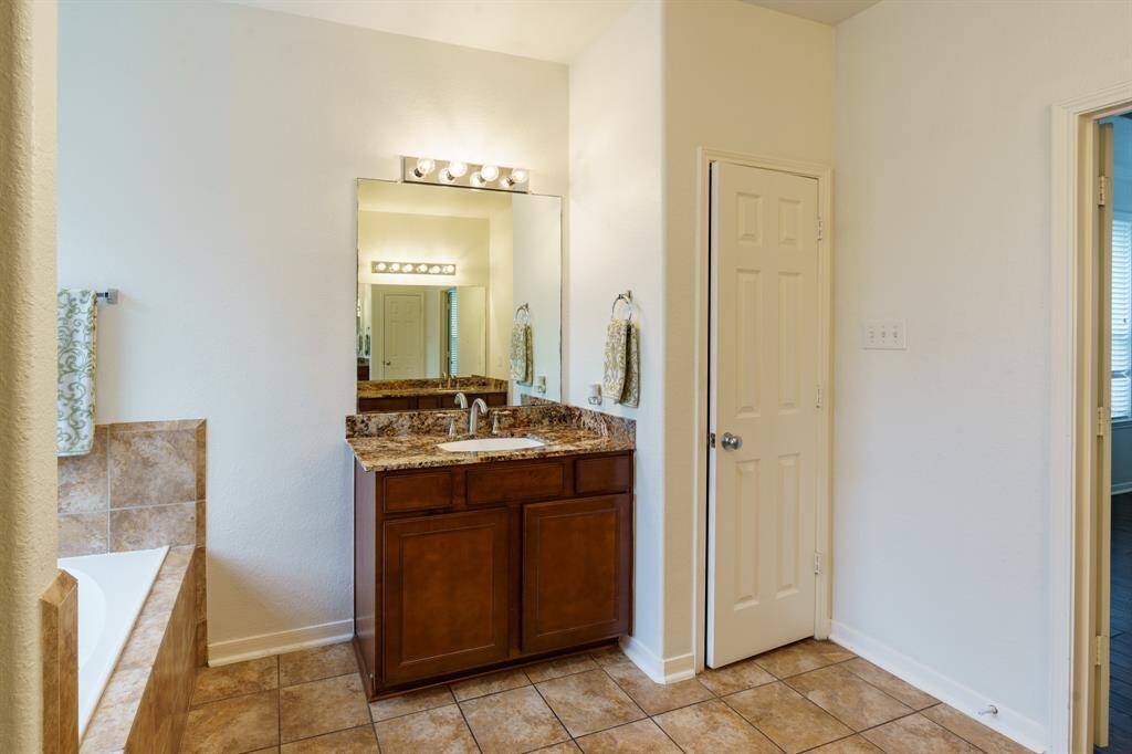 This bathroom features a spacious layout with a walk-in shower and a soaking tub surrounded by neutral tile. It includes a large mirror above a granite countertop with wooden cabinetry, and a window that provides natural light.