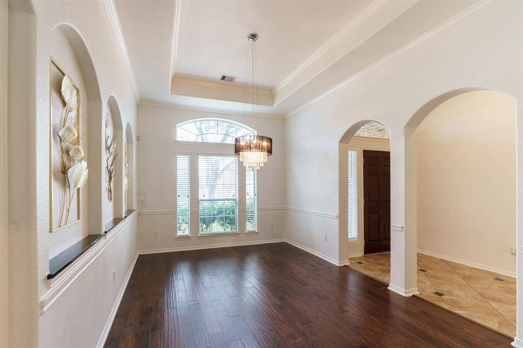 This elegant Dining Room features rich hardwood flooring, a stunning chandelier, and decorative wall niches with gold accents. The space is bright with a high tray ceiling, offering a sophisticated atmosphere perfect for dining or a sitting area.