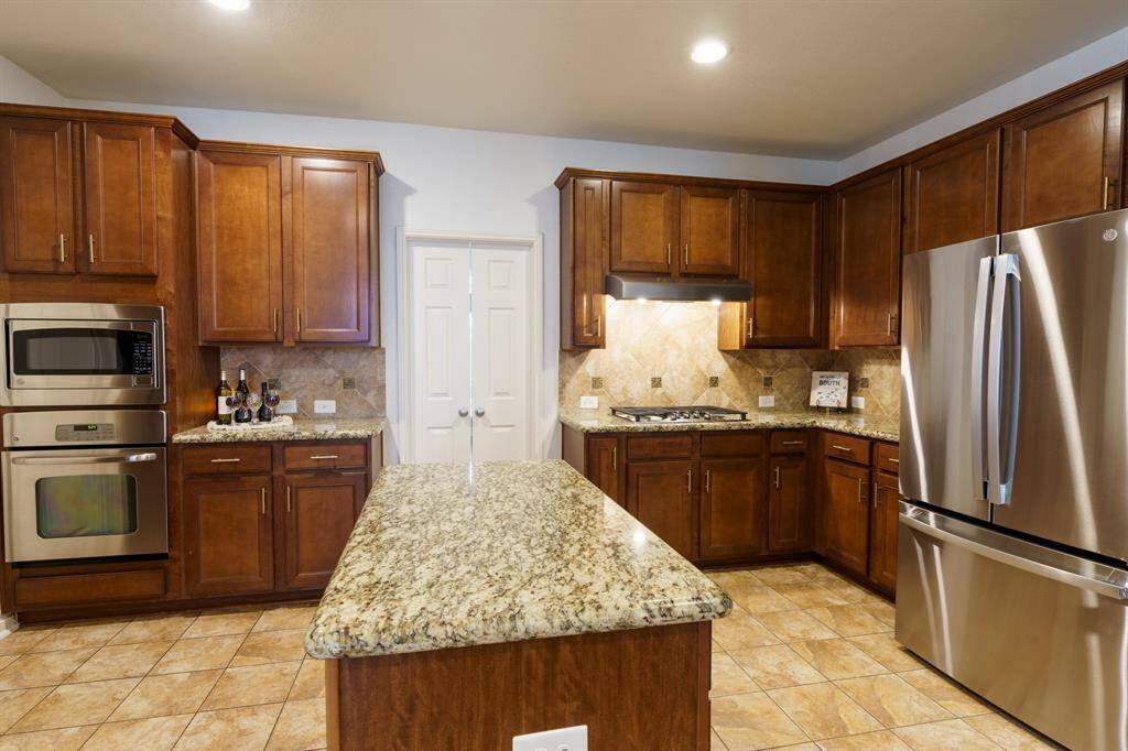 This kitchen features warm wood cabinetry, granite countertops, and a central island. It includes stainless steel appliances, tiled flooring, and arches opening to an adjacent living area, offering a spacious and elegant cooking environment.