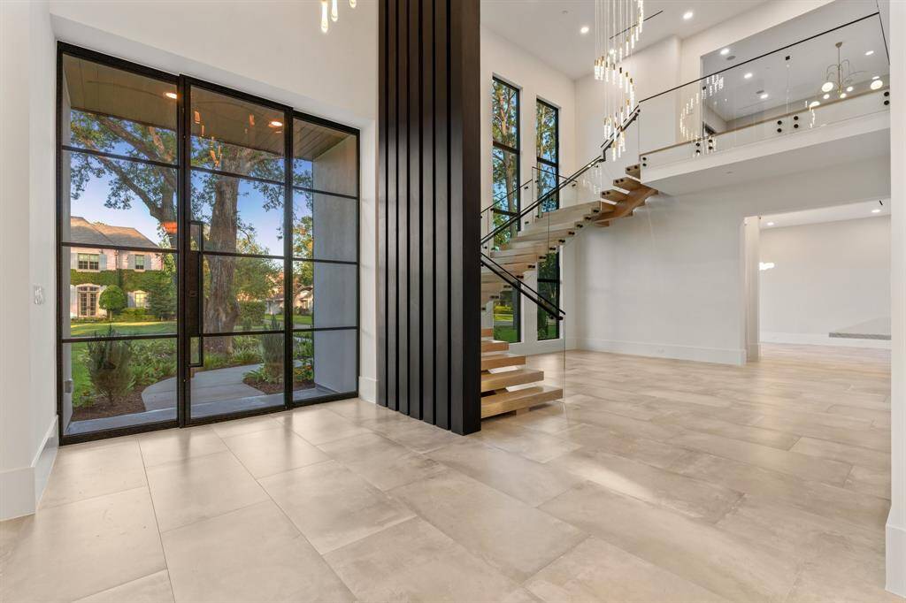 The double-height foyer makes a dramatic first impression with large-format porcelain tile floors, drop-down chandeliers, walls of windows, and a beautiful open staircase with glass handrails.