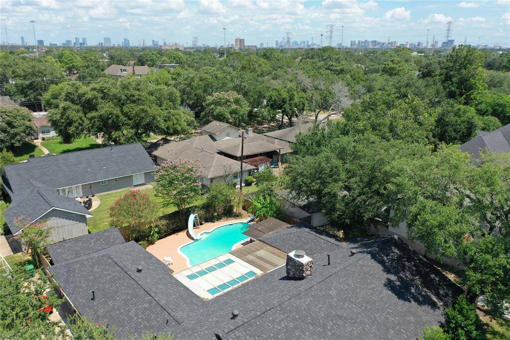 This aerial photo showcases a residential neighborhood with single-family homes surrounded by mature trees. One property features a private swimming pool with surrounding patio area, offering a relaxing outdoor space. The skyline of a city is visible in the distance, suggesting an urban proximity.