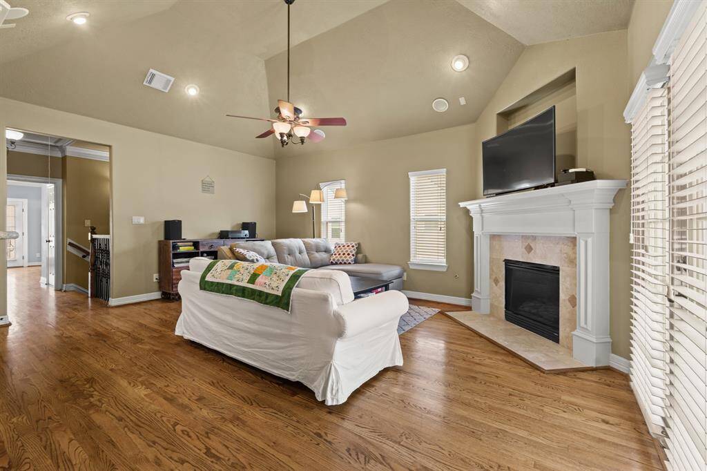 This stunning living room features a vaulted ceiling with a ceiling fan, wood floors, windows with privacy blinds, and a gorgeous fireplace with mantle.