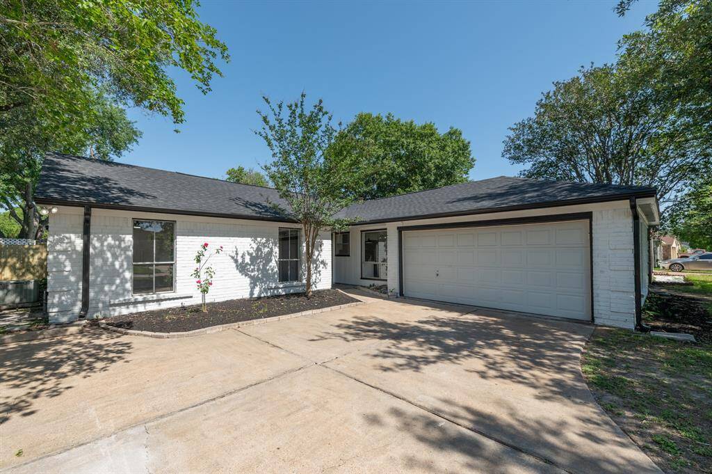 A charming single-story home with a white brick exterior and a dark roof. It features a spacious attached two-car garage and a simple front yard with a small garden bed and mature trees, providing a welcoming and shaded entrance.