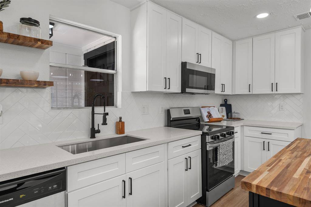 Bright kitchen area with modern white cabinetry, brand new stainless steel dishwasher, and light wood like LVT flooring. Large window offers natural light and views of a lush serene greenery area in the front entrance. The teak wood floating shelves give this kitchen a stylish touch.